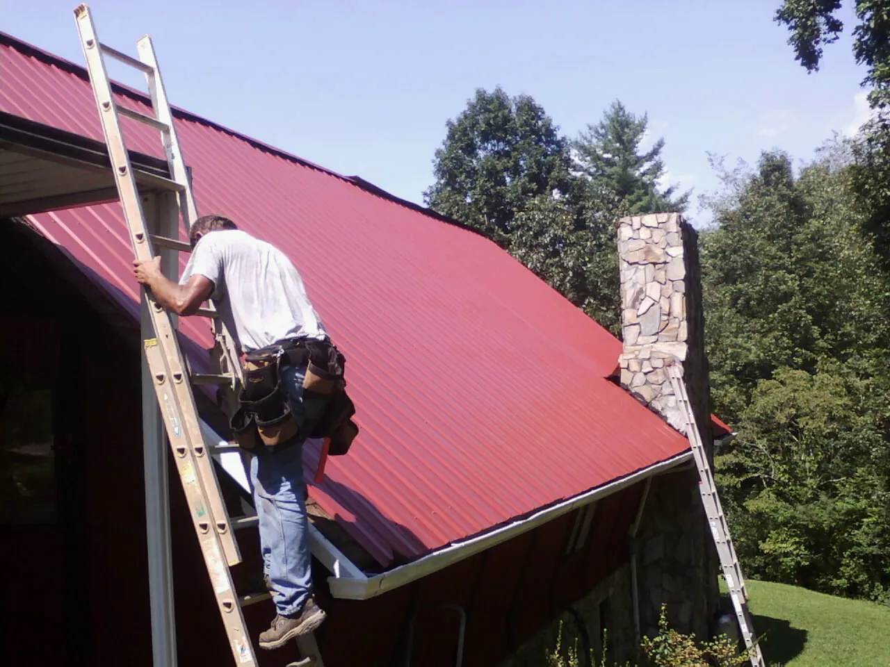 Roofer on a ladder beside a red metal roof and stone chimney — Soffit & Fascia Repair by Roofing Brothers in Felton